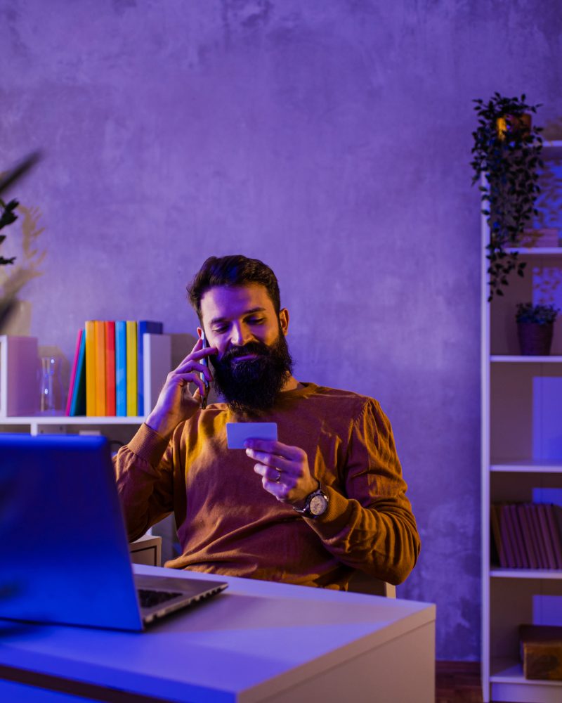 A happy entrepreneur is sitting in a neon blue and purple lighted home office with a credit card.